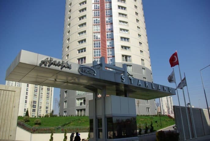 Modern tasarımlı, uzun, dairesel bir konut binası, berrak mavi bir gökyüzünün önünde duruyor. Önünde, Türk bayrağı ve yakınlarda dalgalanan ek bayraklarla "STARLAND" etiketli bir yapı var. Alan peyzajlı ve girişin yakınında yürüyen üç kişi görülüyor.