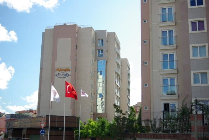 Resimde çok katlı, modern, ten rengi bir apartman kompleksi yer alıyor. Binanın ön cephesinde balkonlar ve büyük pencereler var. Ön planda Türk bayrağı da dahil olmak üzere kırmızı ve beyaz bayraklar var. Gökyüzü birkaç dağınık bulutla açık.