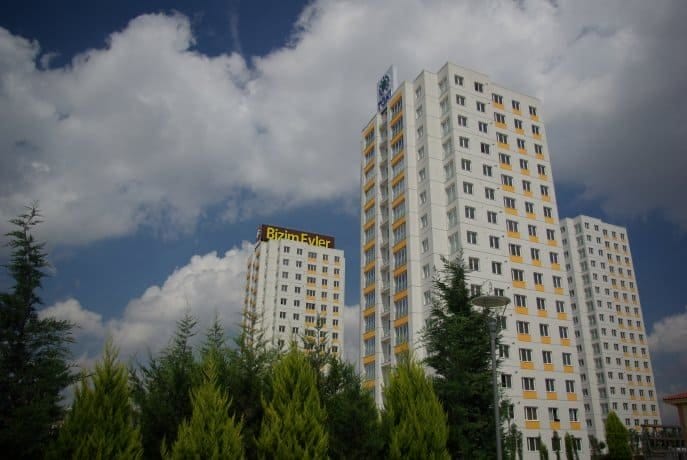 Çok katlı üç yüksek konut binası, bir sıra yeşil ağacın arkasında duruyor. Binaların beyaz ve sarı bir cephesi var ve birinin tepesinde "Bizim Evler" tabelası belirgin bir şekilde yer alıyor. Yukarıdaki gökyüzü kısmen bulutlu.