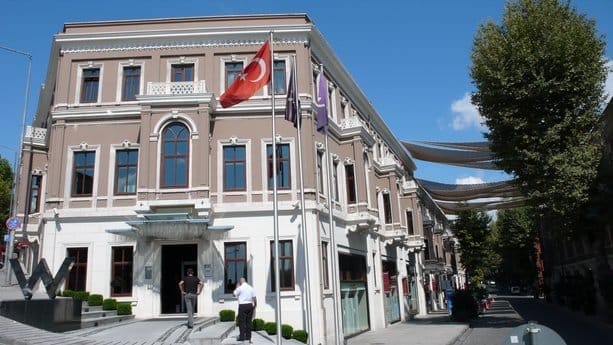 Güneşli bir günde, beyaz çerçeveli ve çok sayıda pencereli büyük, zarif bej bir bina, bir sokağın köşesinde duruyor. Önündeki üç bayrak direğinde Türk bayrağı ve iki bayrak daha var. Girişin yakınında iki kişi var. Geniş bir merdiven binanın ana girişine çıkıyor.