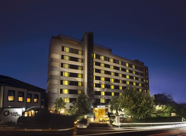 Çok sayıda aydınlatılmış pencereye sahip modern çok katlı bir otelin gece görünümü. Bina ağaçlar ve peyzajla çevrilidir. Ön planda, sokaktan geçen araçlardan gelen ışık izleri sahneye dinamik bir unsur katmaktadır. Gökyüzü karanlıktır ve aydınlatılmış cepheyi vurgular.