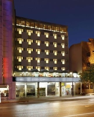 Modern bir cepheye, aydınlatılmış pencerelere ve zemin katında bir McDonald's restoranına sahip yedi katlı bir bina. Önündeki sokak ıslak, binadan ve yakındaki yapılardan gelen ışıkları yansıtıyor ve yakın zamanda yağan yağmuru gösteriyor. Çevrede aydınlatılmış pencereleri olan ek binalar da var.