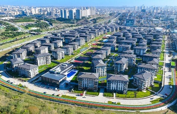 TEMA İSTANBUL'un havadan görünümü, çok sayıda aynı çok katlı apartman binasının bir ızgara düzeninde düzenlendiği modern bir yerleşim alanı. Yeşil çimenler ve park edilmiş arabaların olduğu yollar binaları çevreliyor. Yüksek apartman kuleleri ve şehir manzarası, berrak mavi gökyüzünün altında arka planda.