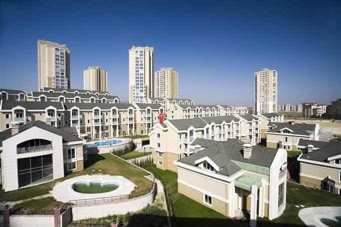 Arka planda yüksek bej kuleleri ve ön planda yeşil çatılı daha küçük bej şehir evleri bulunan çok katlı apartman binalarının bulunduğu bir yerleşim alanı. Manzaranın ortasında küçük, boş bir dairesel yüzme havuzu görülüyor. Yukarıdaki gökyüzü berrak ve mavi.