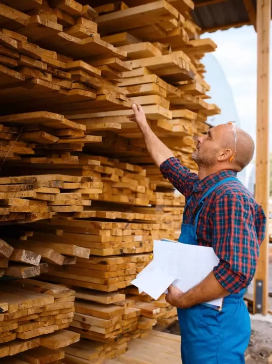 In the About Us section, a person wearing blue overalls and a red plaid shirt is inspecting stacks of cut lumber in an outdoor setting. They hold papers in their left hand and raise their right hand to touch one of the wooden planks. Behind them are piles of similar wooden boards under a shelter.