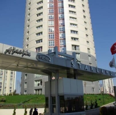 Modern tasarımlı, uzun, dairesel bir konut binası, berrak mavi bir gökyüzünün önünde duruyor. Önünde, Türk bayrağı ve yakınlarda dalgalanan ek bayraklarla "STARLAND" etiketli bir yapı var. Alan peyzajlı ve girişin yakınında yürüyen üç kişi görülüyor.