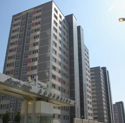 Three high-rise apartment buildings with numerous windows under a clear blue sky. A sign with illegible text is displayed on a structure in the foreground. Streetlights and other buildings are visible in the distance. The setting appears to be urban.