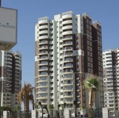 A cluster of modern high-rise apartment buildings stands under a clear blue sky. In the foreground, a large sign reads "Soyak" and "Mavişehir". The buildings have balconies and are surrounded by palm trees. A black fence runs along the bottom of the image.