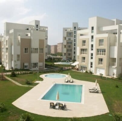 A residential complex featuring several modern, multi-story apartment buildings with balconies. In the center, there are two outdoor swimming pools with lounge chairs and umbrellas. The area is landscaped with grassy lawns and scattered shrubs. Other similar buildings are visible in the background.