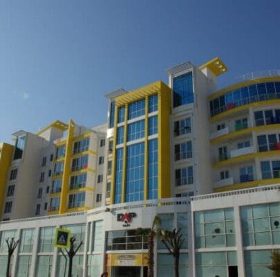 Modern multi-story building with a white facade and vibrant yellow accents. The structure has a mix of glass and solid walls, balconies, and a central glass tower. The ground floor features a glass-walled commercial area with the logo "DAP" at the entrance. Palm trees line the street.