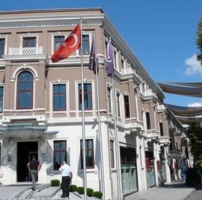 Güneşli bir günde, beyaz çerçeveli ve çok sayıda pencereli büyük, zarif bej bir bina, bir sokağın köşesinde duruyor. Önündeki üç bayrak direğinde Türk bayrağı ve iki bayrak daha var. Girişin yakınında iki kişi var. Geniş bir merdiven binanın ana girişine çıkıyor. Güneşli bir günde, beyaz çerçeveli ve çok sayıda pencereli büyük, zarif bej bir bina, bir sokağın köşesinde duruyor. Önündeki üç bayrak direğinde Türk bayrağı ve iki bayrak daha var. Girişin yakınında iki kişi var. Geniş bir merdiven binanın ana girişine çıkıyor.