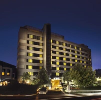 A tall, modern hotel building with illuminated windows at night. The hotel has multiple floors and is surrounded by green trees. A well-lit entrance is visible, with a single car parked outside. Adjacent to the hotel is a smaller, older-style building also lit up, all set against a deep blue night sky.