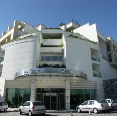 Modern tasarımlı, yeşilliklerle süslenmiş balkonlara sahip çok katlı bir otel. Girişinde “Royal Asarlik Beach” yazan gümüş bir tabela bulunan kavisli bir cephe var. Önünde birkaç araba park edilmiş ve gökyüzü açık, bazı dağınık bulutlar var. Modern tasarımlı, yeşilliklerle süslenmiş balkonlara sahip çok katlı bir otel. Girişinde "Royal Asarlik Beach" yazan gümüş bir tabela bulunan kavisli bir cephe var. Önünde birkaç araba park edilmiş ve gökyüzü açık, bazı dağınık bulutlar var.