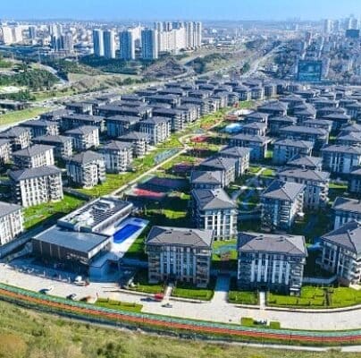 TEMA İSTANBUL'un havadan görünümü, çok sayıda aynı çok katlı apartman binasının bir ızgara düzeninde düzenlendiği modern bir yerleşim alanı. Yeşil çimenler ve park edilmiş arabaların olduğu yollar binaları çevreliyor. Yüksek apartman kuleleri ve şehir manzarası, berrak mavi gökyüzünün altında arka planda.