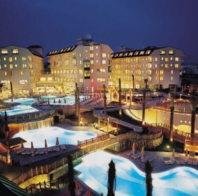 A luxurious resort illuminated at night with several interconnected, brightly lit swimming pools. Surrounding the pools are lounge chairs, umbrellas, and palm trees. The resort buildings have multiple floors with glowing windows, indicating occupancy. The sky is dark, enhancing the serene ambiance.