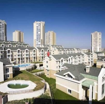 Arka planda yüksek bej kuleleri ve ön planda yeşil çatılı daha küçük bej şehir evleri bulunan çok katlı apartman binalarının bulunduğu bir yerleşim alanı. Manzaranın ortasında küçük, boş bir dairesel yüzme havuzu görülüyor. Yukarıdaki gökyüzü berrak ve mavi.