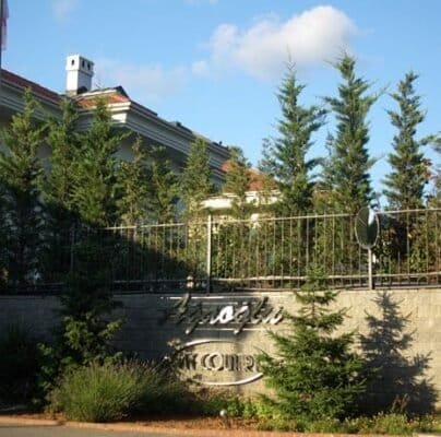 A two-story white building with a red-tiled roof is partially obscured by tall evergreen trees and a reflective metal fence. A gate beside the building features an ornate sign with decorative text. The scene is bathed in warm sunlight under a blue sky with a few scattered clouds.