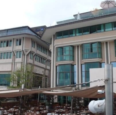 Ahşap masa ve sandalyelerin bulunduğu açık oturma alanı kısmen bej kanopilerle kaplıdır. Arkasında, çok sayıda uzun pencere, cam panel ve bej renkli dış duvarlara sahip, modern mimari tasarıma sahip büyük, çok katlı bir bina bulunmaktadır. Ağaçlar ve yeşillikler görülebilir. Ahşap masa ve sandalyelerin bulunduğu açık oturma alanı kısmen bej kanopilerle kaplıdır. Arkasında, çok sayıda uzun pencere, cam panel ve bej renkli dış duvarlara sahip, modern mimari tasarıma sahip büyük, çok katlı bir bina bulunmaktadır. Ağaçlar ve yeşillikler görülebilir.