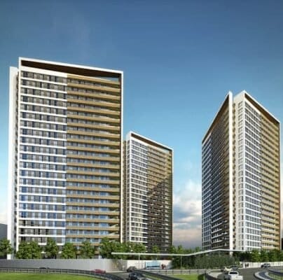 A modern architectural scene featuring three tall, rectangular residential buildings with numerous windows and balconies. The buildings, crafted with wood accents, are surrounded by trees and a road in the foreground, with a partly cloudy sky in the background.