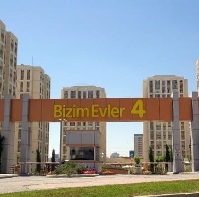 “Bizim Evler 4” yazan büyük bir tabelanın bulunduğu bir konut kompleksi girişi. Kapılı giriş, modern bir mimari stilde çok sayıda yüksek apartman binasıyla çevrilidir. Ağaçlar ve peyzajlı yeşillikler, arka planda berrak mavi bir gökyüzü ile alanı süslüyor. "Bizim Evler 4" yazan büyük bir tabelanın bulunduğu bir konut kompleksi girişi. Kapılı giriş, modern bir mimari stilde çok sayıda yüksek apartman binasıyla çevrilidir. Ağaçlar ve peyzajlı yeşillikler, arka planda berrak mavi bir gökyüzü ile alanı süslüyor.