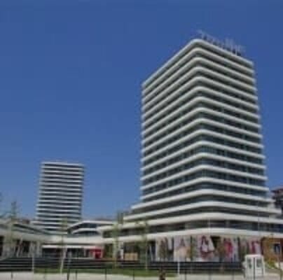 A modern building complex featuring a tall, multi-story structure with horizontal white balcony lines and large glass windows. Nearby, a shorter building of similar design stands against a clear blue sky. A landscaped area with trees and a billboard are visible in the foreground.