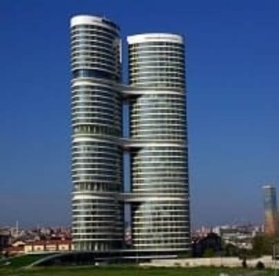 A modern, cylindrical twin tower rises against a clear blue sky. Each tower is connected by three bridges at different heights. The buildings feature numerous windows, forming a sleek and reflective facade. In the background, another set of tall skyscrapers is visible. The surrounding area includes smaller buildings and greenery.