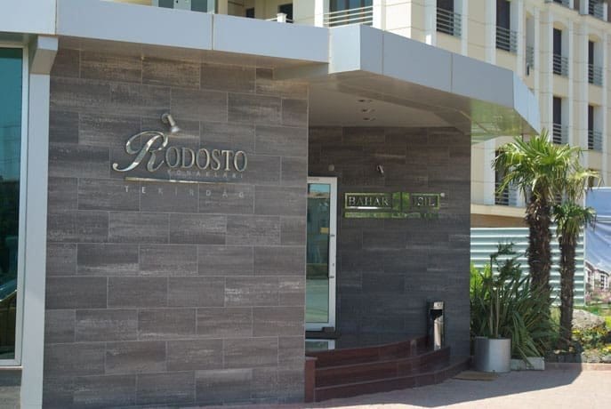 Entrance of a modern building named "RODOSTO". The facade features dark grey tiles and glass. A door with a green sign reading "Bahar Etil" is visible. Outdoor plants, including a small palm tree, decorate the entrance area. The building has multiple floors with light-colored exterior walls and balconies.
