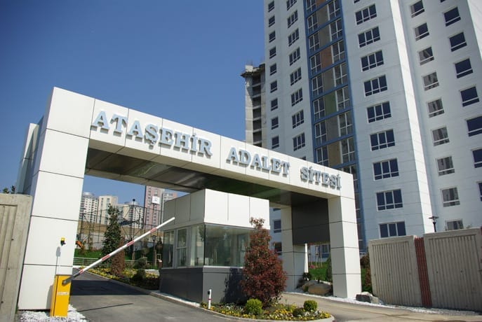 "Ataşehir Adalet Sitesi" olarak etiketlenen modern bir konut kompleksi girişi. Girişte kapılı bir güvenlik kulübesi ve kompleksin adının yazılı olduğu bir tabela bulunmaktadır. Arka planda, çok sayıda pencereye sahip yüksek, beyaz apartman binaları, berrak mavi bir gökyüzünün altında yer almaktadır.