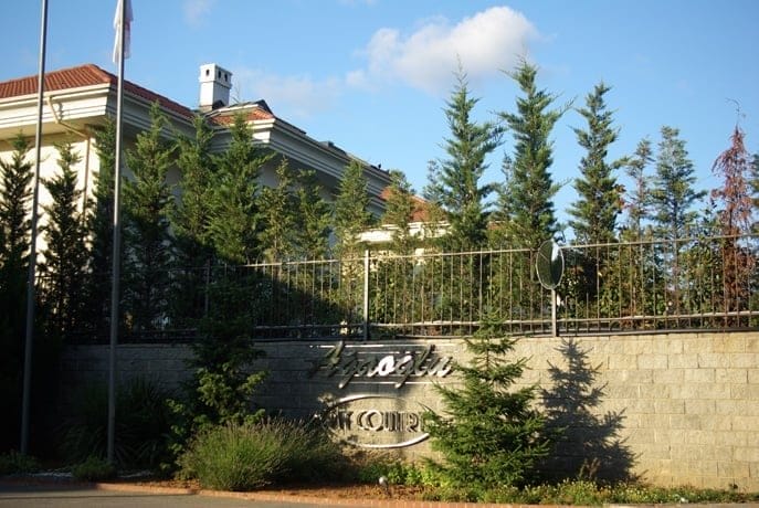 Uzun, herdem yeşil ağaçlarla kısmen kaplı bir binanın ve demir çitli yüksek bir tuğla duvarın sokak seviyesinden görünümü. Güneş ışığı, üzerinde metin bulunan metalik bir tabela bulunan duvara gölgeler düşürüyor. Binanın önünde küçük bir çimenlik alan ve bir bayrak direği görülüyor.