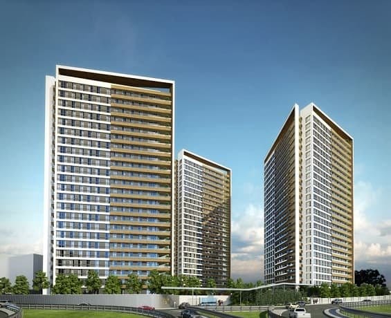 Yansıtıcı cam cephelere sahip üç modern yüksek apartman binası, mavi gökyüzünün altında yan yana duruyor ve her biri çok sayıda balkona sahip. Ön planda araçların olduğu bir yol var ve binaların tabanında ağaçlar ve yeşillikler var ve girişlerinin ahşap vurgularını tamamlıyor.