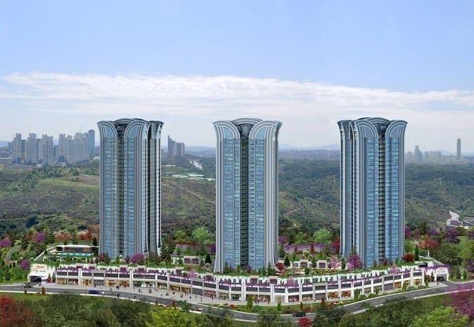 Three tall, modern residential towers with a unique curved design stand amidst greenery and hills in the background. The complex includes a row of commercial spaces at the base, flanked by lush trees and colorful landscaping. Wooden doors add a rustic charm, while the city skyline is visible under a clear sky.