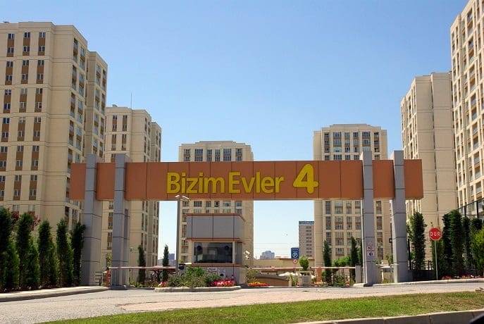 The image shows the entrance to a residential complex named "Bizim Evler 4." The complex features several cream-colored high-rise buildings with balconies. The entrance has a large sign with the complex's name in yellow letters on a beige and orange structure. Tall trees line the pathway leading to the buildings.