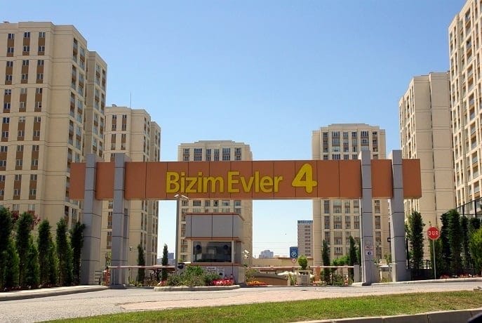 "Bizim Evler 4" yazan büyük bir tabelanın bulunduğu bir konut kompleksi girişi. Kapılı giriş, modern bir mimari stilde çok sayıda yüksek apartman binasıyla çevrilidir. Ağaçlar ve peyzajlı yeşillikler, arka planda berrak mavi bir gökyüzü ile alanı süslüyor.