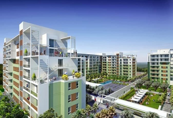 A modern residential complex with several multi-story apartment buildings featuring balconies and rooftop terraces. The central area includes landscaped gardens, a pool, and recreational amenities. The buildings are light green and beige, and the sky is clear and blue.
