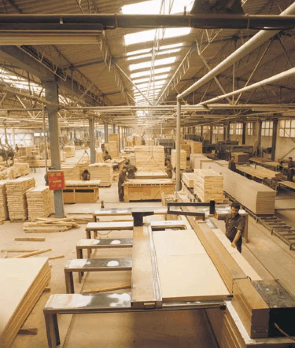 A large, industrial woodworking factory with stacks of wooden panels and planks organized throughout the space. Workers are operating machinery and working on various woodworking tasks. The ceiling has exposed beams, and the area is well-lit with rows of overhead lights. For inquiries, contact us today.