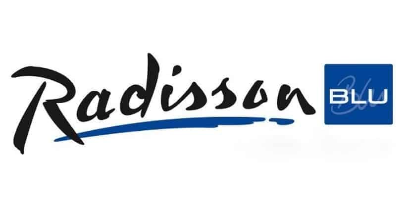 Resimde Radisson Blu logosu gösteriliyor. "Radisson" kelimesi, mavi alt çizgi fırça darbesiyle şık siyah bir el yazısıyla yazılmış. Sağda, mavi bir karede beyaz büyük harflerle "BLU" kelimesi yer alıyor.