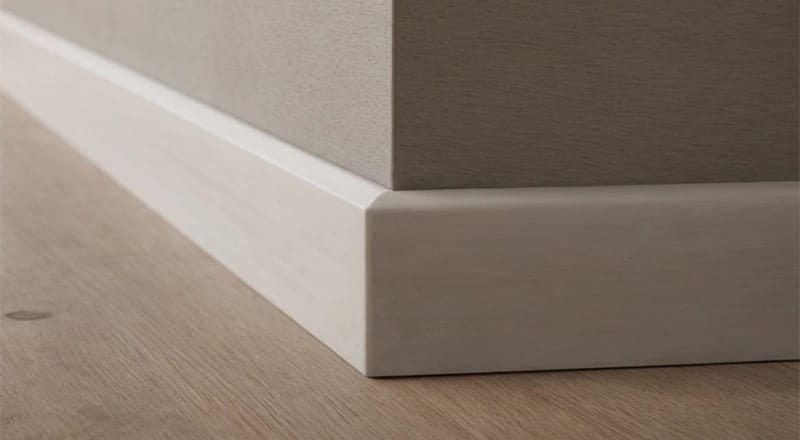 Close-up of a beige wall corner with white skirting boards meeting a light wood floor. The baseboards are sleek and modern, fitting neatly at the junction of the wall and floor, providing a clean and polished look to the interior space.