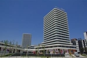 Modern, çok katlı bir ofis binası, berrak mavi bir gökyüzüne karşı dimdik duruyor. Yapının dış kısmında katmanlı bir etki yaratan şık, beyaz yatay çizgiler yer alıyor. Ana binayı benzer tasarıma sahip ek, daha kısa yapılar çevreliyor. Alan ağaçlar ve yeşilliklerle düzenlenmiş.
