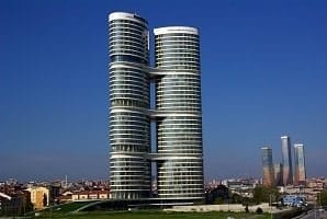 A modern, cylindrical twin tower rises against a clear blue sky. Each tower is connected by three bridges at different heights. The buildings feature numerous windows, forming a sleek and reflective facade. In the background, another set of tall skyscrapers is visible. The surrounding area includes smaller buildings and greenery.